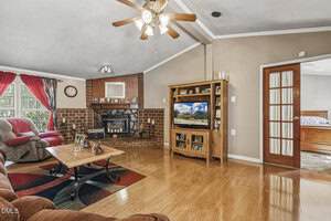 A living room with wood flooring, brown sofas, a brick fireplace, a wooden entertainment center with a TV, a coffee table, and a ceiling fan. A door with glass panes leads to a bedroom. Large windows let in natural light.