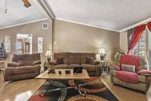 A living room with a brown sofa, matching armchair, and recliner, all positioned around a wooden coffee table with framed photos. Two lamps are on side tables; a window with red curtains lets in natural light.