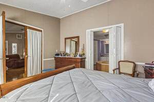 A bedroom with a wooden bed, dresser with a mirror, and an upholstered chair. Double doors lead to a bathroom with a bathtub, and another set of doors opens to an adjacent room with seating.