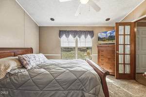 A bedroom with a wooden sleigh bed, gray bedding, a purple pillow, a wood dresser with a TV on top, and a window with blinds and a valance. A ceiling fan is above, and French doors lead to another room.