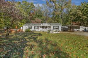 A single-story white house with a front porch sits in a large yard covered with scattered autumn leaves, surrounded by trees. A wooden deck with an umbrella is on the left, and a metal carport is on the right.