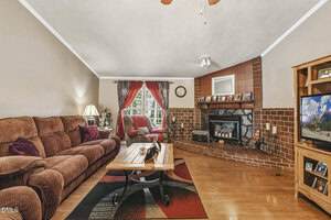 Living room with brown sectional sofa, wooden coffee table, brick fireplace with mantel, wood flooring, TV on a stand, and a window with red curtains. Decor includes a table lamp, photos, and a ceiling fan.