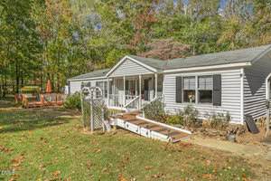 Single-story white house with a covered front porch, ramp, and flower beds. Wooden deck with patio umbrella is visible to the left. Yard has scattered leaves and is surrounded by trees with fall foliage.