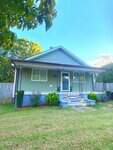 A single-story house at 306 Chavasse Avenue in Henderson features light gray siding, a small covered front porch, blue steps, and white trim. A few shrubs and plants accent the grassy yard, with a wooden fence in the background.