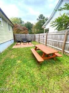 A backyard with a wooden picnic table on grass, a paved patio area with a table and chairs, and a wooden fence separating the yard from neighboring houses.