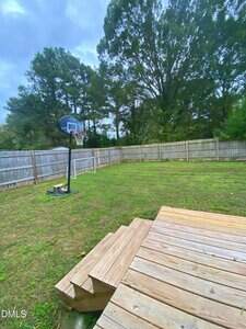 A backyard with a wooden deck, several wooden steps leading to the grass, a basketball hoop on the left, and a wooden privacy fence enclosing the area. Tall trees are visible behind the fence under a cloudy sky.