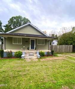Single-story house with light green siding, white trim, and a covered front porch. There are bushes in front, concrete steps leading to the porch, and a driveway with a wooden fence on the right.