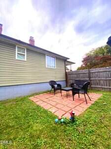 A small backyard patio with red square pavers, two black wicker chairs, a matching table, and a wooden fence. Lawn surrounds the patio, and a few bocce balls and a drink can are on the grass nearby.