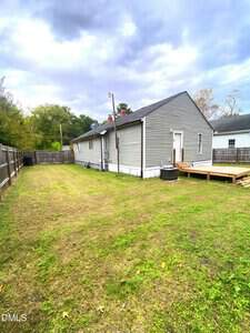 A small gray house with white trim sits in a fenced backyard with a grassy lawn, wooden deck, and central air unit. The sky is overcast and neighboring houses are visible on either side.