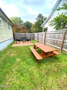 A fenced backyard with a grassy lawn, a wooden picnic table in the foreground, and a paved patio area with a table and four chairs near the back fence. Trees and another house are visible in the background.