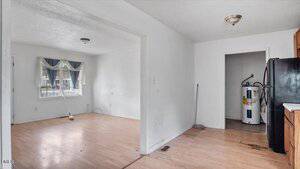 A mostly empty room with light wood flooring, white walls, a window with blue curtains, a broom, and cleaning supplies. The adjoining kitchen has dark cabinets, a black fridge, and a water heater visible in a utility area.