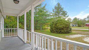 View from a covered wooden porch with white railings, looking out onto a grassy yard with a large bush, trees, a dirt driveway, and a red building in the background under a blue sky.
