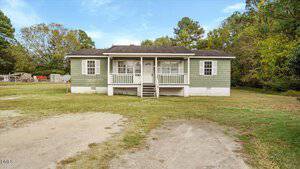 Single-story green house with white trim and a covered front porch, set on a grassy lot with trees in the background and a dirt driveway in the foreground.