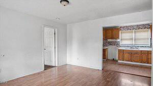 An empty room with white walls and light wood flooring, connected to a small kitchen with wood cabinets and a tile backsplash. A door is open to another room on the left. Ceiling light fixture is visible.