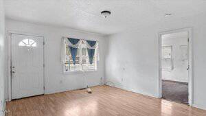 Unfurnished room with white walls, light wood flooring, a front door, a window with blue and white curtains, a broom leaning against the wall, and an open doorway leading to another room.