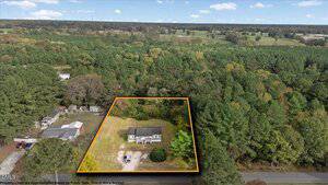 Aerial view of a house situated on a rectangular, outlined lot surrounded by trees and neighboring properties, with a road in the foreground and fields and woodland in the background.