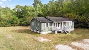 A small green house with white trim and a front porch, sitting on a large grassy lot with trees in the background under a blue sky with some clouds.