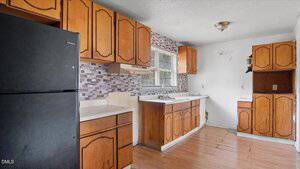 A kitchen with wood cabinets, a black refrigerator, a stove hood, and a double sink under a window. The backsplash features a mosaic tile pattern, and the floor is wood. Some wall areas show visible wear.