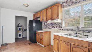 A kitchen with wood cabinets, a black refrigerator, a double sink under a window, and a tile backsplash. An adjacent room has a water heater and a broom. The floor has some stains near the refrigerator.