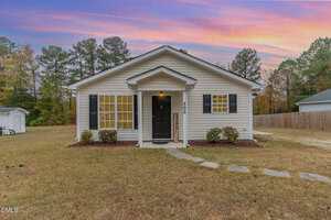 A small, single-story white house with black shutters stands on a grassy yard at sunset on 505 Cotton Avenue in Creedmoor. Stone pavers lead to a black door, two lights, and a vertical welcome sign, with trees lining the background.