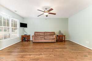 A living room with hardwood floors, a brown sofa against a pale green wall, two wooden side tables, a small wall-mounted TV, and a ceiling fan with lights. Large windows let in natural light from the left.