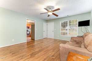 A living room with light green walls, wood flooring, a ceiling fan with lights, a wall-mounted TV, a brown sofa, and a small side table. An open doorway leads to a kitchen with stainless steel appliances.