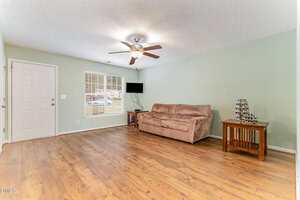 A living room with wood flooring, a beige couch, a small wooden side table with a wine rack, a wall-mounted TV, and a ceiling fan with lights. The walls are light green and there is a window and a white door.