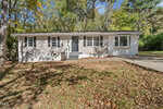 Charming single-story house at 70 Lynnbank Estates Road in Kittrell features light gray siding, white trim, a dark front door, and multiple windows. Nestled among trees, the yard is blanketed with fallen leaves and a driveway sits to the right.