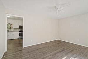 A room with light-colored walls, wood-look flooring, a white ceiling fan, and an open doorway leading to a kitchen with white cabinets and stainless steel appliances.