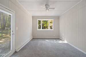 A small, empty room with light grey carpet, beige vertical panel walls, a white ceiling with a ceiling fan, a door with blinds on the left, and three windows showing green trees outside.