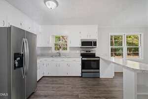 A kitchen with white cabinets, stainless steel appliances including a refrigerator, oven, and microwave, a double sink, marble-patterned countertops, and large windows overlooking greenery. The floor is dark wood.