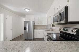 A modern kitchen with white cabinets, stainless steel appliances including a refrigerator, oven, and microwave, light-colored countertops, and wood-look flooring under a ceiling light.