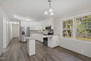 A kitchen with white cabinets, stainless steel appliances, marble-pattern countertops, and wood-look flooring. There is a large window by the dining area, letting in natural light and showing trees outside.