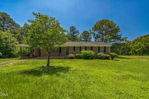 Charming single-story brick home at 605 Sagefield Way, Henderson, features a covered carport on the left, lush green grass, a front yard tree, and tall trees in the background beneath a clear blue sky.