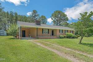 Single-story brick house with a covered carport, central chimney, and multiple windows on the front. The yard is grassy with a few shrubs and trees, and there are tall pine trees in the background.