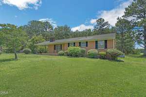 Single-story brick house with black shutters and a large front lawn, surrounded by trimmed bushes and trees, under a partly cloudy sky.