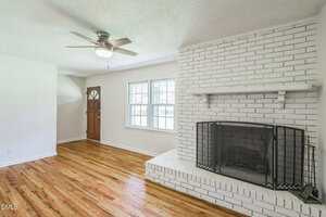 A living room with hardwood floors, white brick fireplace with a black screen, ceiling fan with light, and a wooden front door next to two windows letting in natural light.