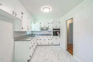 A kitchen with white cabinets, silver handles, granite countertops, and a stainless steel oven. The floor is tiled in white with gray veins, and there is a circular ceiling light. An open doorway leads to another room.