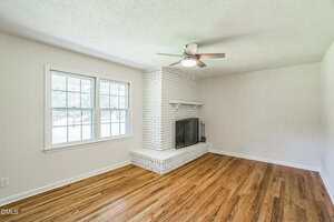 A bright living room with hardwood floors, a white brick fireplace with a black fireguard, a ceiling fan with a light, and a large window letting in natural light. Walls are painted light beige.