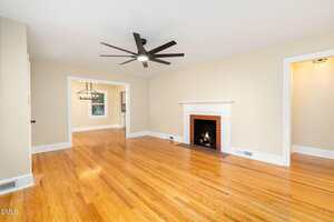 A spacious living room with polished hardwood floors, beige walls, a white fireplace with a lit fire, a modern ceiling fan, and white trim around doorways and baseboards. An adjoining room is visible in the background.