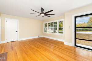 A bright, empty room with light tan walls, hardwood floors, a large black ceiling fan, a white door, a set of three windows, and a glass door leading outside.
