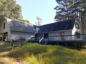 A two-story gray house with a sloped roof, large wraparound porch, wooden ramp, and steps. The yard has tall grass, a split-rail fence, and is surrounded by trees under a clear sky.