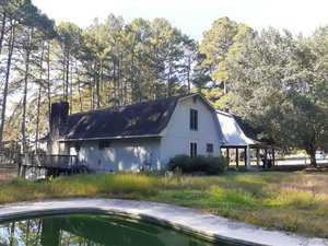 A two-story, barn-style house with gray siding and a black roof is surrounded by tall pine trees. There is a covered carport area and an in-ground pool with green water in the foreground.