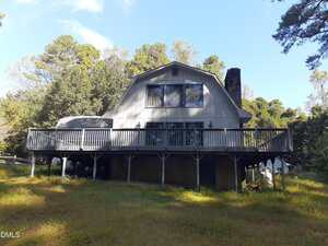 A two-story house with a gray exterior and a large wraparound wooden deck, elevated on stilts, set among trees and grass under a clear blue sky.