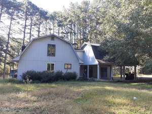 A two-story house at 1576 Munns Road, Creedmoor, features gray siding, a barn-style roof on the left wing, and a covered porch on the right, surrounded by tall pine trees and overgrown grass in the yard.