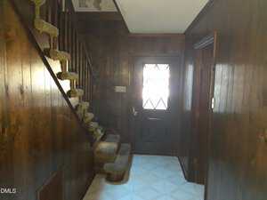Interior hallway with dark wood-paneled walls, a staircase with carpeted steps and railing on the left, a door with a diamond-patterned window in the center, and tiled floors. Light enters through the door window.