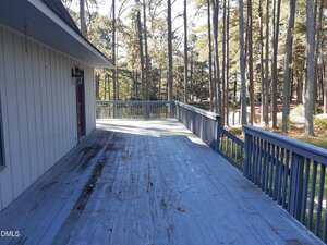 A large wooden deck with worn blue-gray paint extends from a house, bordered by a simple railing and overlooking a yard with many tall pine trees. Sunlight filters through the trees, casting shadows on the deck.