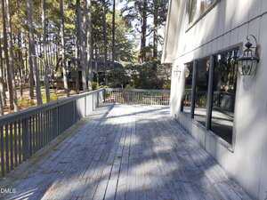 A large wooden deck with weathered blue-gray paint is attached to a house with gray siding and large windows. The deck overlooks a wooded area with tall pine trees and nearby houses.