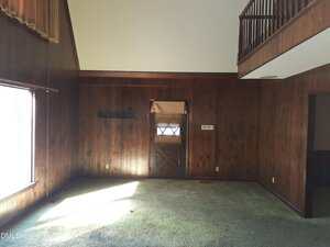 A room with dark wood-paneled walls, greenish carpet, a window letting in sunlight, a door with a small window and blinds, and an upper balcony railing on the right above part of the room.