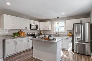 Modern kitchen with white cabinets, stainless steel appliances, granite countertops, center island, and wood-look flooring. A window above the sink lets in natural light. Decorative plants and kitchen items are on the counters.
